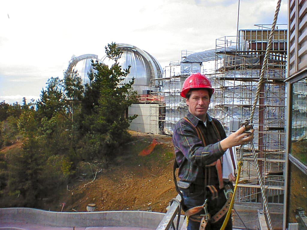 A construction worker in a red hard hat uses safety gear while standing on a scaffold near a building under construction; the impressive dome structure in the background highlights advanced architecture design.