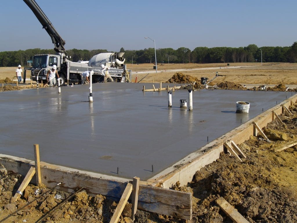 Freshly poured concrete foundation slab at a construction site, featuring exposed plumbing pipes and equipment in the background—a crucial step in residential engineering and architecture design.