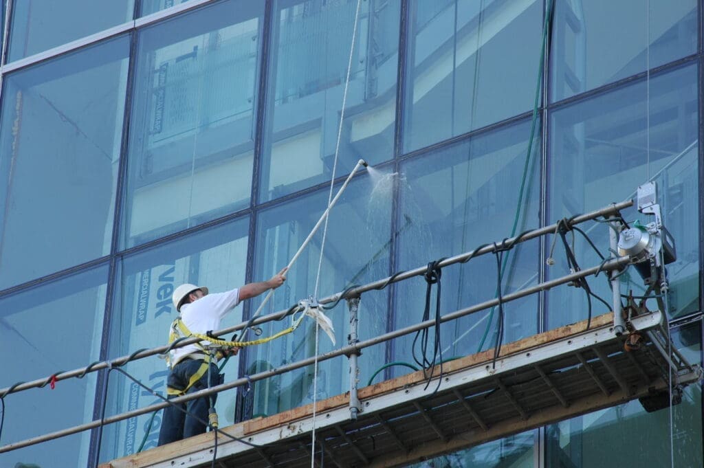 A worker wearing a safety harness cleans the windows of a glass building with a long pole from a suspended platform, maintaining the curtainwall facades that highlight innovative architecture design.