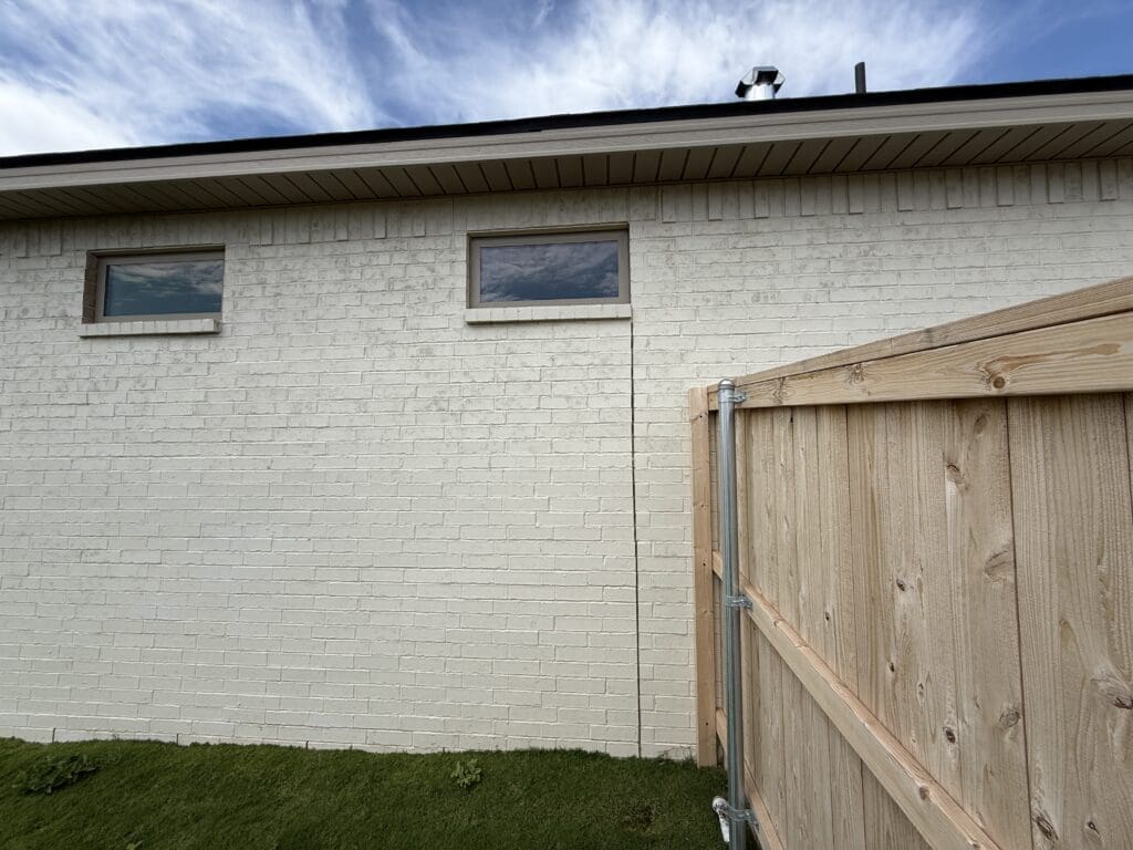A white brick exterior wall with two small rectangular windows, a wooden fence gate, and green artificial grass in the foreground under a partly cloudy sky showcases thoughtful architecture design.