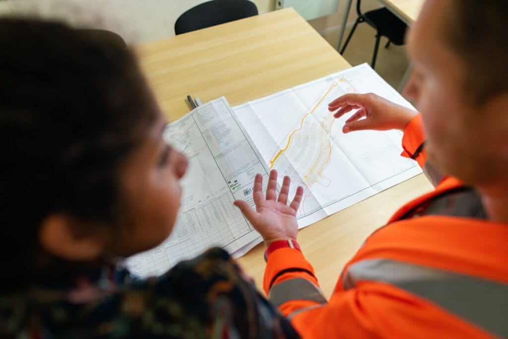 Two people review a technical map or blueprint at a desk; one points to details while discussing engineering rules of thumb, while the other listens attentively.