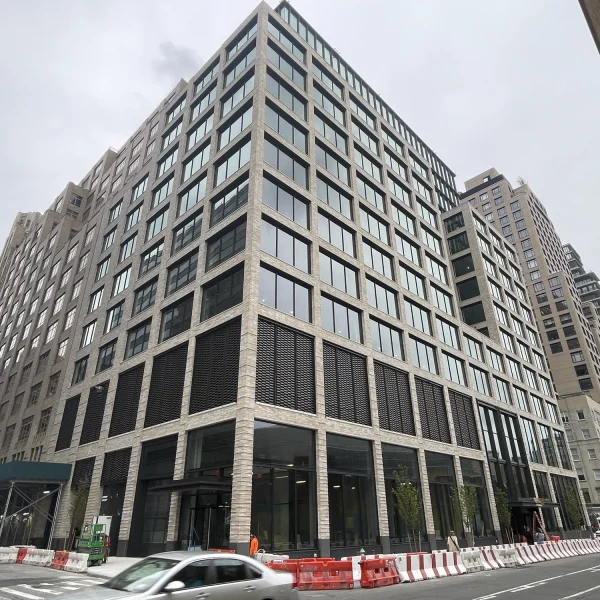 A large modern office building with striking architecture design features many windows on a city street, bordered by construction barriers and a passing white car.