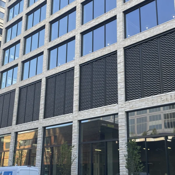 Modern office building with large glass windows and curtainwall facades, featuring louvered panels on the lower floors. Situated beside another contemporary structure, with a white van parked in front.