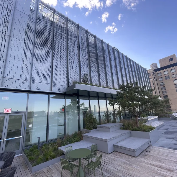 Modern rooftop terrace with seating, greenery, and a metal curtainwall facade on one building; adjacent architecture design is visible under a partly cloudy sky.
